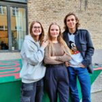 Tre elever, to piger og en dreng, smiler til kameraet i Gefions skolegård. Bag dem ses den gule skolebygning og et rød og grønt bordtennisbord.