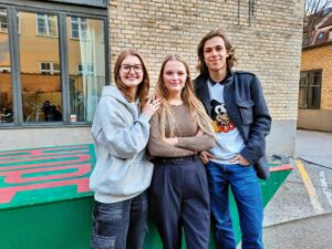 Tre elever, to piger og en dreng, smiler til kameraet i Gefions skolegård. Bag dem ses den gule skolebygning og et rød og grønt bordtennisbord.