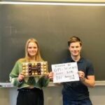 To elever, en pige og en dreng, viser præmierne fra Gefionmesterskaberne i Science. Pigen holder en æske chokolade, og drengen holder et papir med teksten "Vinder af GM i Science 2022, kemi". Bag dem ses en tavle.