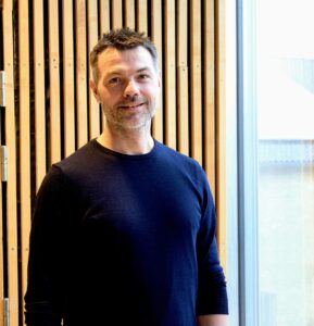 Portræt af Vicerektor Benjamin Haahr Bach. Baggrunden består af et vindue i højre side og en væg af lodrette, lyse stykker træ i venstre side.