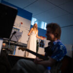 En dreng med en guitar i forgrunden. En pige i hvid kjole synger på scenen, i baggrunden.