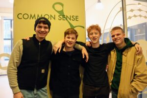 Fire gefionelever står ved siden af hinanden og holder om hinanden med en arm om sidemandens hals. Thomas (t.v.), Anders (mf.), Bertram (mf.) og Sigurd (th.). På væggen bag dem ses teksten "common sense".