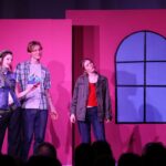 Gefions elevproducerede musical 2024: Mean Girls. Scene med lyserød baggrund. Scene med en dreng og to piger, der ser ud mod højre.