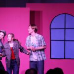Gefions elevproducerede musical 2024: Mean Girls. Scene med lyserød baggrund. En dreng og t piger taler sammen på scenen, ser ud på publikum.