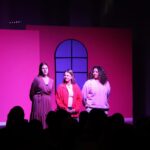 Gefions elevproducerede musical 2024: Mean Girls. Scene med lyserød baggrund. Tre piger på scenen, ser ud på publikum. Tre piger er de tre instruktører, der præsenterer stykket.