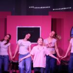Gefions elevproducerede musical 2024: Mean Girls. Scene med lyserød baggrund. En pige synger, andre piger danser i baggrunden.