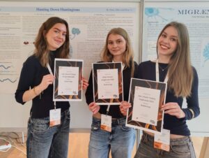Tre elever står ved en tavle med deres plakat om Huntingtons sygdom. Hver elev holder et diplom op foran sig. DrugHunters 2025.