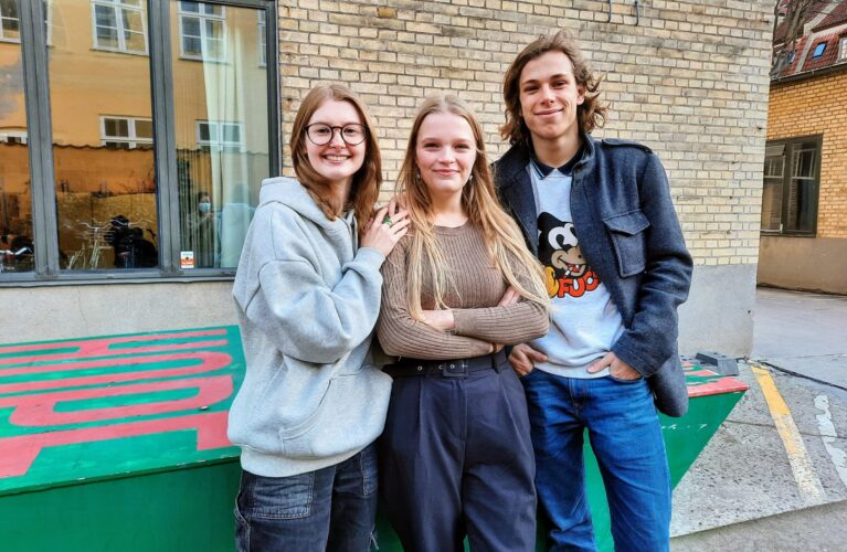 Tre elever, to piger og en dreng, smiler til kameraet i Gefions skolegård. Bag dem ses den gule skolebygning og et rød og grønt bordtennisbord.