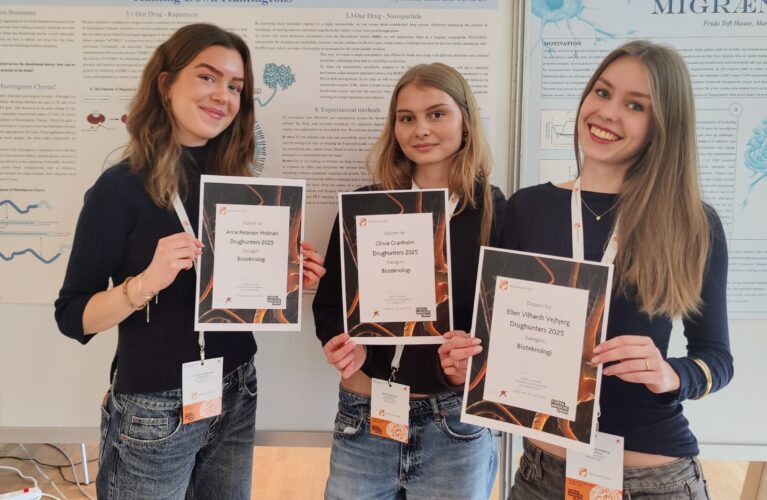 Tre elever står ved en tavle med deres plakat om Huntingtons sygdom. Hver elev holder et diplom op foran sig. DrugHunters 2025.