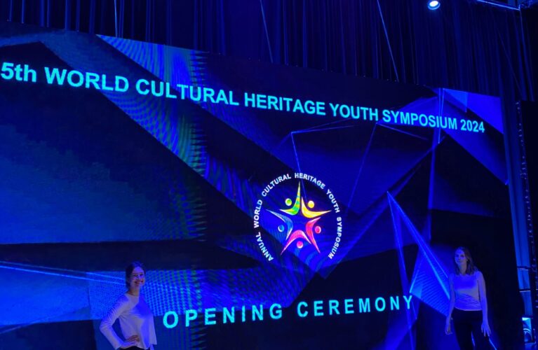 To piger foran en væg belyst med mørkeblåt lys med teksten: "5th world cultural heritage youth symposium 2024. Opening ceremony. Helenic cultural and educational club for Unesco"