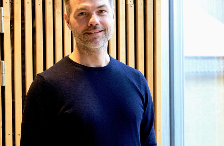 Portræt af Vicerektor Benjamin Haahr Bach. Baggrunden består af et vindue i højre side og en væg af lodrette, lyse stykker træ i venstre side.